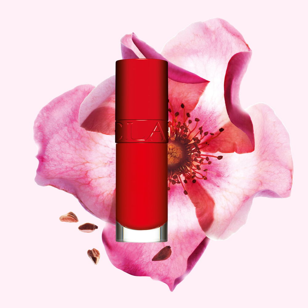 Packshot av Clarins Lip Comfort Oil i Iconic Red, placerad bredvid en nyponros blomma p&aring; en svart-r&ouml;d bakgrund.