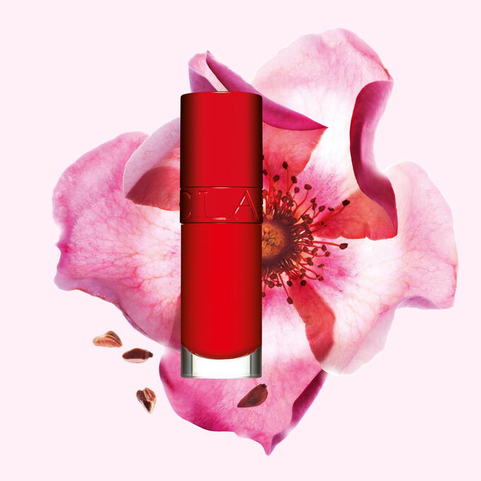 Packshot av Clarins Lip Comfort Oil i Iconic Red, placerad bredvid en nyponros blomma p&aring; en svart-r&ouml;d bakgrund.