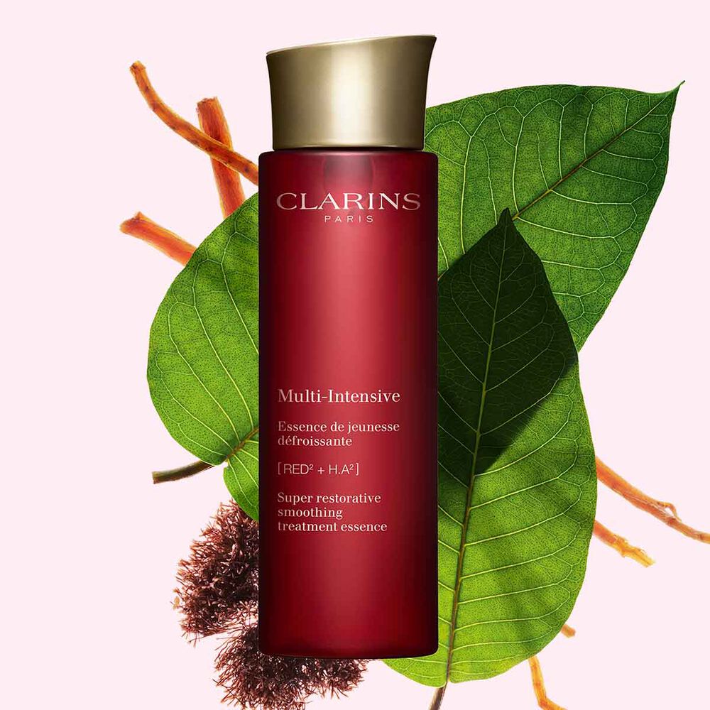 Packshot av Clarins Super Restorative Treatment Essence nära rödalg, harunganablad och ginsengrot mot en rosa bakgrund.