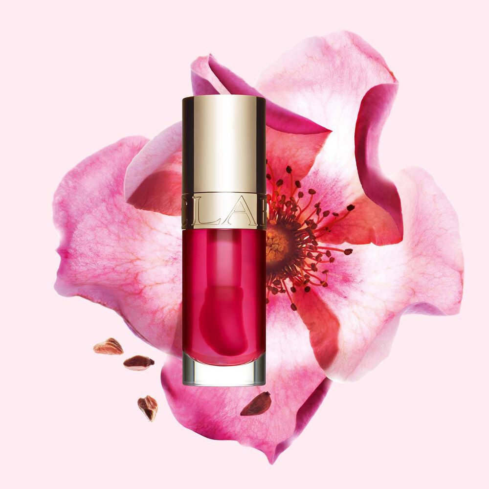 Packshot av Clarins Lip Comfort Oil i Pitaya, placerad bredvid en nyponros p&aring; en ljus rosa bakgrund.