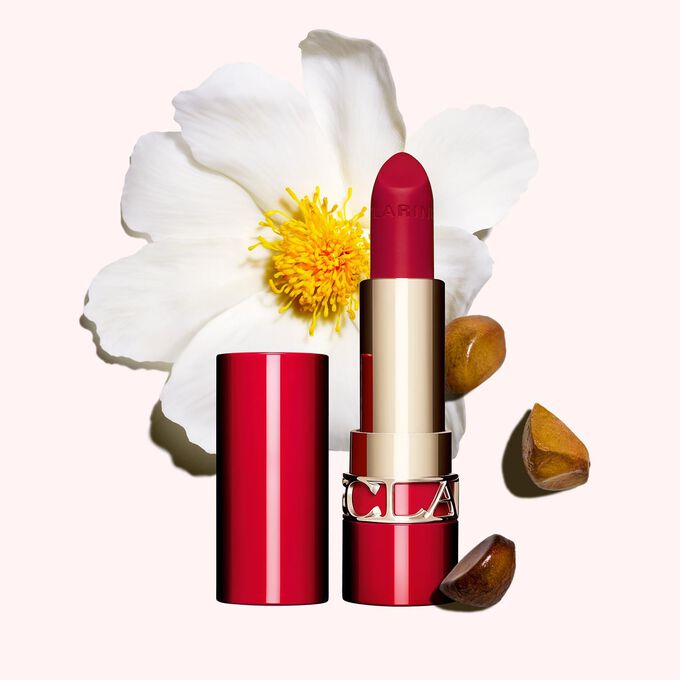 Packshot av Clarins matta läppstift Joli Rouge i nyansen 742 V, placerat bredvid en kamelia blomma och sheafrön på en rosa bakgrund.