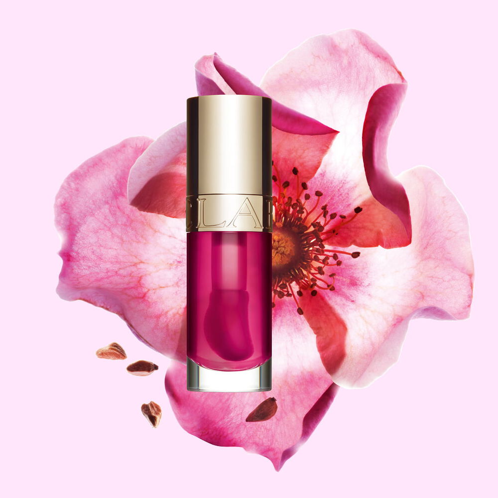Packshot av Clarins Lip Comfort Oil i Raspberry, placerad bredvid en nyponros p&aring; en ljus persikof&auml;rgad bakgrund.