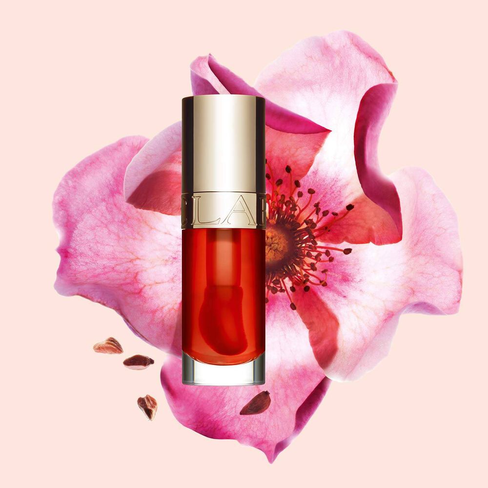 Packshot av Clarins Lip Comfort Oil i Apricot, placerad bredvid en nyponros p&aring; en ljus orange-pesikof&auml;rgad bakgrund.
