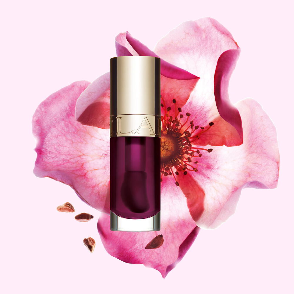 Packshot av Clarins Lip Comfort Oil i Plum, placerad bredvid en nyponros p&aring; en ljus rosa bakgrund.