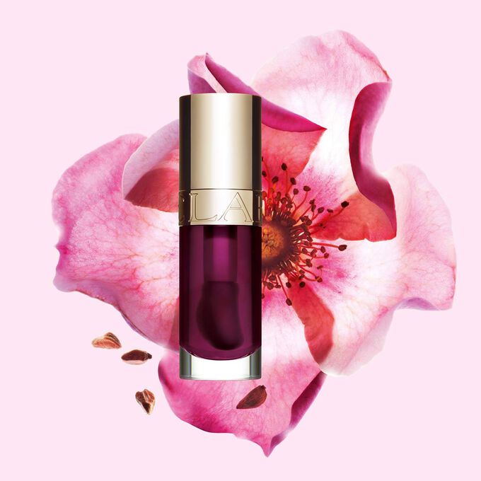Packshot av Clarins Lip Comfort Oil i Plum, placerad bredvid en nyponros p&aring; en ljus rosa bakgrund.