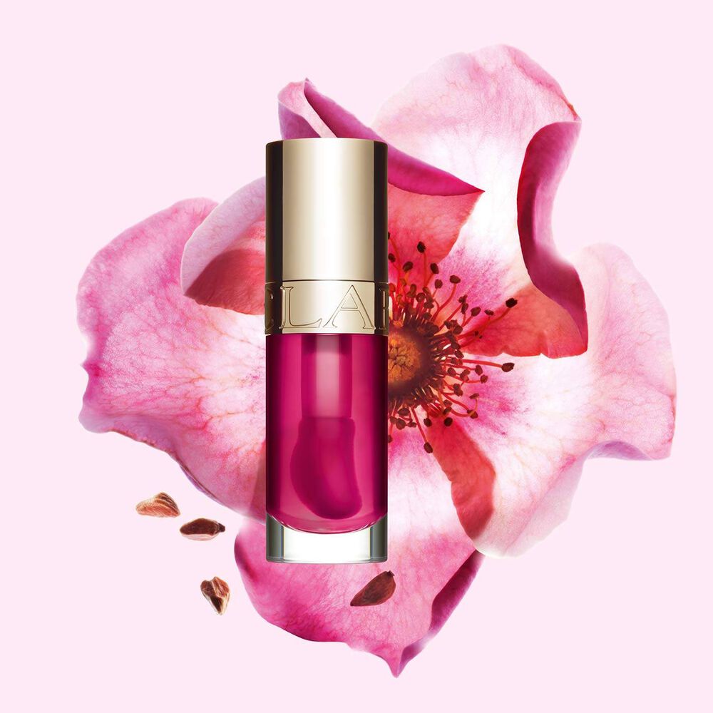 Packshot av Clarins Lip Comfort Oil i Raspberry, placerad bredvid en nyponros på en ljus persikofärgad bakgrund.
